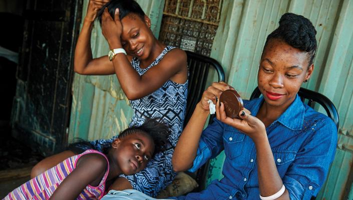 two young women, one fixing her makeup and the other fixing her hair, smile as a younger girl looks on