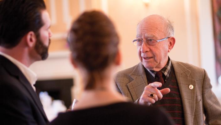 Clive Shiff, wearing a shirt, tie, and jacket speaks with a man and woman whose backs are to the camera