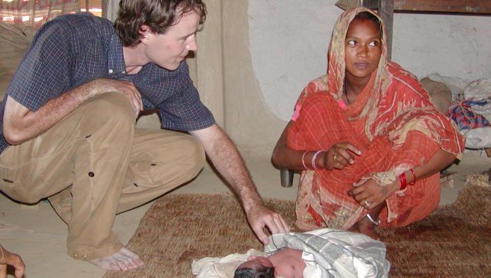 A researcher squats on the floor to examine a newborn infant; the child's mother, in a red sari, sits next to the infant.