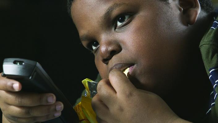 A young boy snacks on chips with a TV remote in his hand