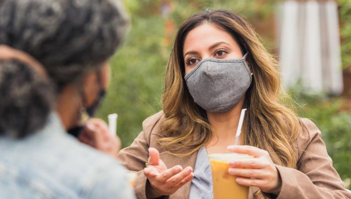 A woman wearing a mask speaks and gestures to a friend