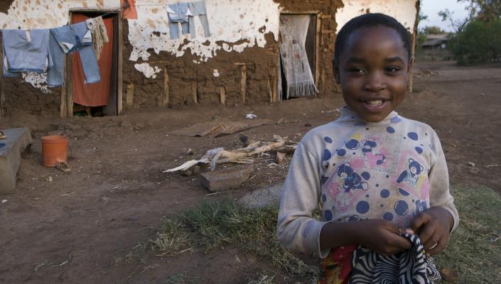 A child wearing a spotted shirt with teddy bears on itin her yard in Tanzania; behind her is her house and a clothesline with clothing hanging on it.