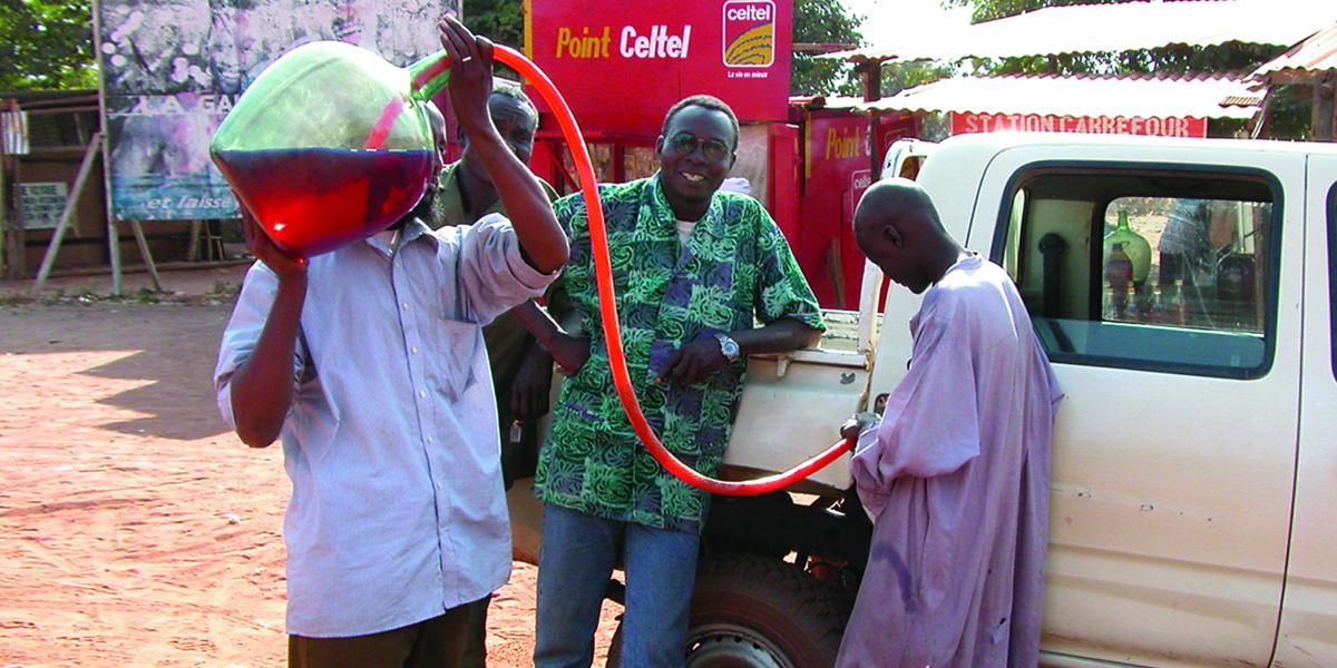 3 men in Chad fill their white pickup's gas tank by siphoning fuel from a glass bottle