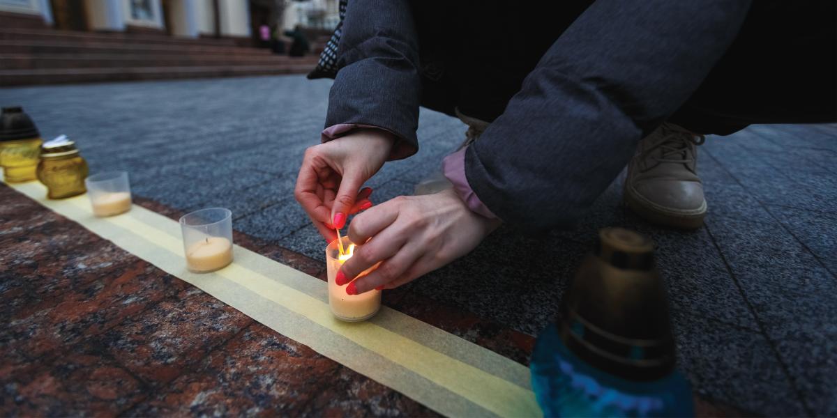 a person kneels to light a candle in a row of candle holders lined up on the street.