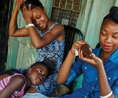 two young women, one fixing her makeup and the other fixing her hair, smile as a younger girl looks on