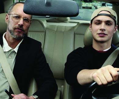 a teen wearing a backward baseball cap sits behind the steering wheel; his father sits in the passenger seat.