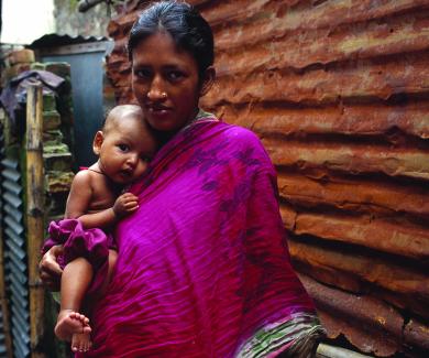  mother in Bangladesh wearing a magenta scarf holds her infant on her hip