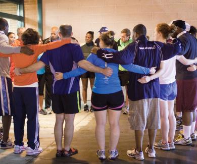 a circle of about 15 people in athletic gear stand in a circle with their arms around each other